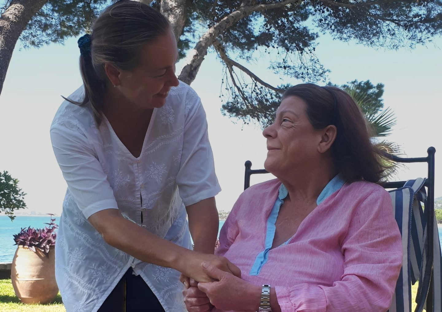 Manuela Dratschew hält die Hände einer sitzenden Frau in einem Garten am Meer auf Mallorca